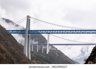 Wide scenic composition of two massive suspension bridges spanning the legendary Tiger Leaping Gorge area over the Jinsha River in Yunnan, China. Framed by a dark, rugged rock face on the right, the vibrant blue steel railway bridge cuts a bold horizontal line across the misty canyon. Far below and behind it, a sleek highway suspension bridge connects the distant foggy peaks. The image perfectly illustrates the immense scale of modern Chinese transport networks built into some of the world's most formidable terrain.