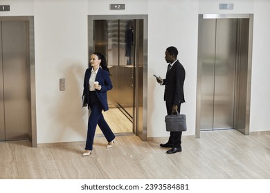 Wide shot of African American businessman holding phone and bag looking at Caucasian business lady exiting elevator in office building - Powered by Shutterstock