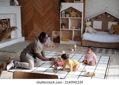 Wide shot of African American father sitting on floor in cozy living room at home having fun playing with his twin babies - Powered by Shutterstock