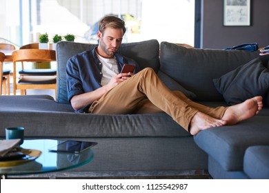 Wide Shot Of An Attractive Caucasian Man Sitting On A Generous Sofa In A Modern Home, While Texting On His Latest Mobile Phone With His Feet Up On The Couch And Relaxing.