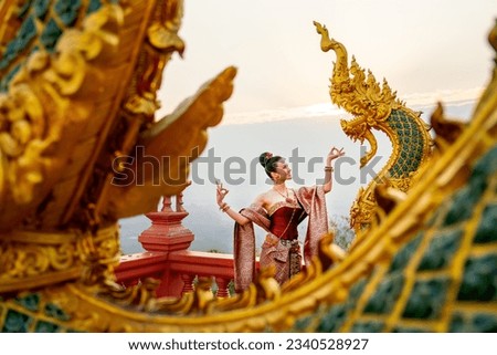 Wide shot of beautiful Asian woman with Thai traditional dress stand with Thai dance action in front of the naga  sculpture in temple area with morning light.