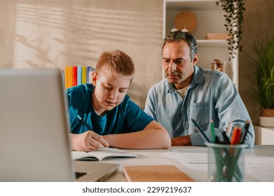 Wide shot of a blond teenage boy sitting at the table at home behind his laptop and writing his homework. His father sitting next to him and looking down at the notebook. Spending weekend studying. - Powered by Shutterstock