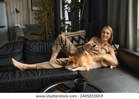 Wide shot of cheerful woman lying on sofa with her dog changing tv channels with remote control