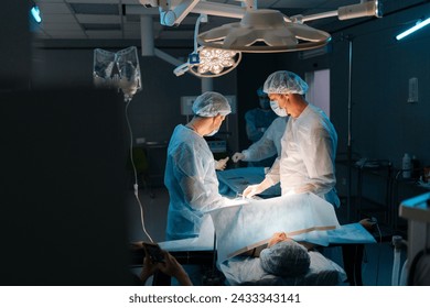 Wide shot of cooperating team of surgeons processing surgical operation in dark operating room modern hospital emergency department. Doctors in sterile gloves, surgical uniforms and masks. - Powered by Shutterstock