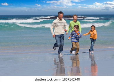 Wide Shot Of A Couple Running With Their Children Along The Shore Of A Beach With Waves In The Background.