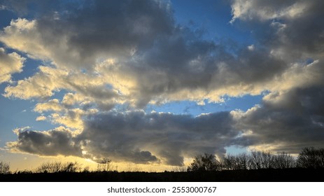 A wide shot depicts a mostly cloudy sky at sunset.  Gray and dark clouds dominate, with lighter, yellowish clouds visible near the horizon. Silhouetted trees are at the bottom. - Powered by Shutterstock