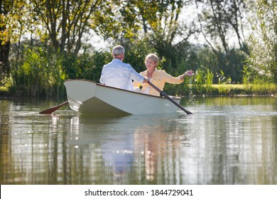 Wide shot of an elderly man rowing a boat over a lake surrounded by trees with his wife watching him.