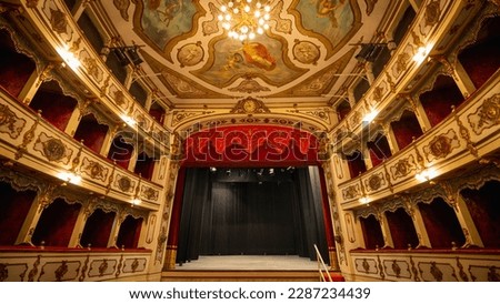 Wide shot of an Empty Elegant Classic Theatre with Big Stage and Red Velvet Curtains. Well-lit Opera House with Beautiful Mural Paintings Ready to Recieve Audience for a Play or Ballet Show