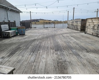 A Wide Shot Of An Empty Wooden Patio Area In The Winter. There Are Strings Of Lights Hung Over The Outside Dining Area.