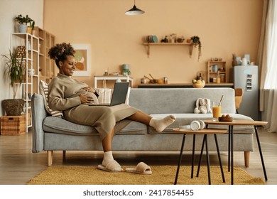 Wide shot of happy pregnant African American woman lying back on big couch in living room with laptop on lap while embracing her round belly - Powered by Shutterstock