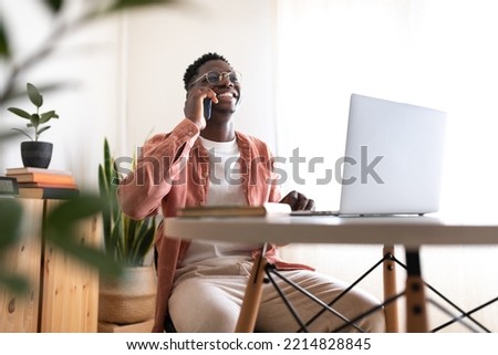 Wide shot of happy, smiling African American black young man talking on the phone while working at home office.