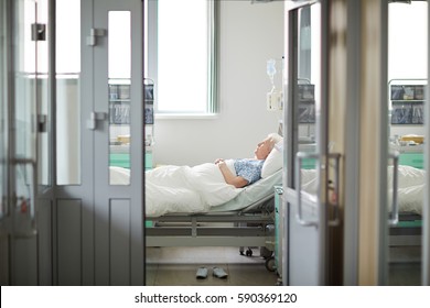 Wide Shot Image Of Doors Opened To Sterile Light Hospital Ward With Sad Lonely Old Woman Lying On Bed In The Room