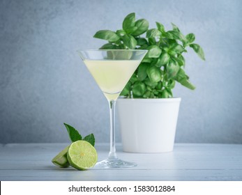 A Wide Shot Of Lime Martini In A Cocktail Glass Near Lime And Mint And A Basil Plant In A White Pot