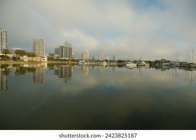 Wide shot Looking north from Demens Landing Park over smooth water with reflections towards city scape of St. Petersburg, Florida .  Sailbots in marina with Cloudy blue and white sky with light fog.
					