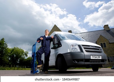 A Wide Shot Of A Man In Coveralls Standing And Posing Between A Hand Truck And A White Work Van.