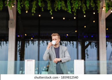 Wide shot of middle aged man leaning on balcony railing, looking away thoughtfully and drinking coffee