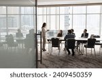 Wide shot of multiethnic business team talking at meeting table in office conference room, brainstorming in co-working space with large window. Project leader woman talking to colleagues