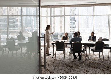 Wide shot of multiethnic business team talking at meeting table in office conference room, brainstorming in co-working space with large window. Project leader woman talking to colleagues - Powered by Shutterstock