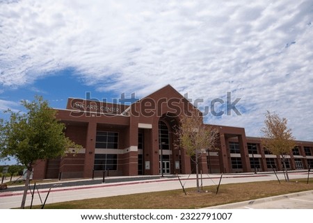 A wide shot of a newly constructed performing arts center brick building on a partly cloudy summer day with newly planted trees in the foreground.
