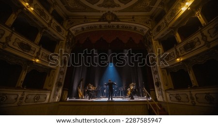 Wide shot of an Orchestra on a Classic Theatre Stage: Professional Conductor Directing Symphony Orchestra with Performers Playing Violins, Cellos, and Trumpets During Music Concert. Audience's POV
