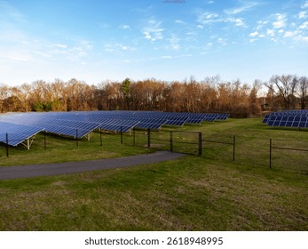 A wide shot of a solar panel array on a grassy field. The panels are arranged in rows and columns, with a fence surrounding the area. Trees are visible in the background under a blue sky