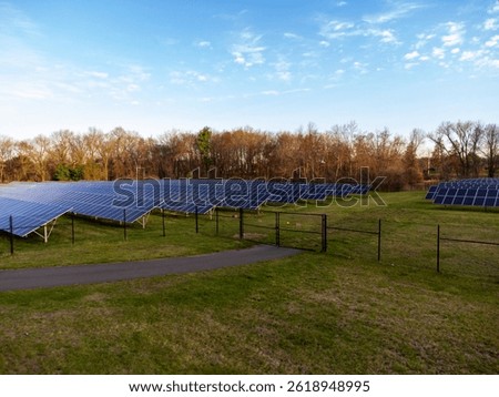 A wide shot of a solar panel array on a grassy field. The panels are arranged in rows and columns, with a fence surrounding the area. Trees are visible in the background under a blue sky