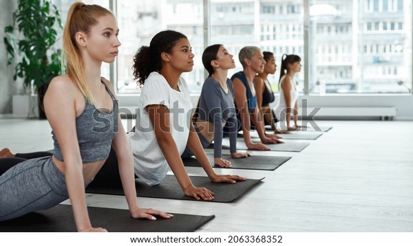 Wide shot of sporty diverse multiethnic women in\
sportswear train practice yoga in fitness club. Active multiracial\
females meditate do sports exercise workout together on stretching\
pilates class.