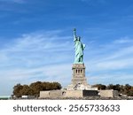 Wide shot of the Statue of Liberty on Liberty Island, framed by trees and a clear blue sky, capturing its monumental presence.