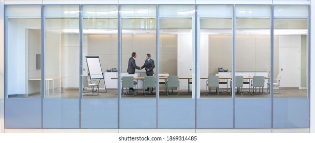 Wide Shot Through An Office Window With Business Men Shaking Hands In The Meeting Room.