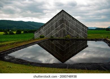A wide shot of a triangular barn with reflection in the water surrounded by country side views and overcast grey clouds