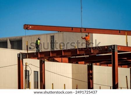 A wide shot of two construction worker directing placement of a large steel beam that is atttached to a crane 