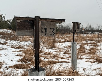 A wide shot of two rustic, weathered wooden bird boxes or nesting houses numbered 13 and 5 on metal posts, standing in a snow-covered field of dry grass under an overcast sky.