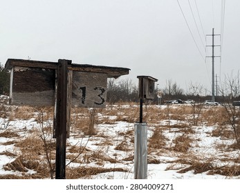 A wide shot of two rustic, weathered wooden bird boxes or nesting houses numbered 13 and 5 on metal posts, standing in a snow-covered field of dry grass under an overcast sky.
