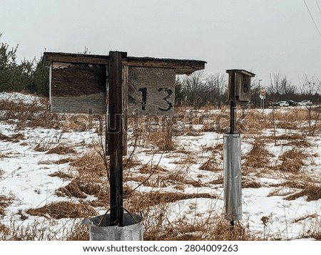 A wide shot of two rustic, weathered wooden bird boxes or nesting houses numbered 13 and 5 on metal posts, standing in a snow-covered field of dry grass under an overcast sky.