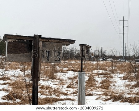 A wide shot of two rustic, weathered wooden bird boxes or nesting houses numbered 13 and 5 on metal posts, standing in a snow-covered field of dry grass under an overcast sky.