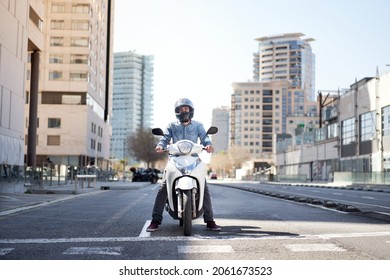 Wide shot of a young motorcyclist stopped at a traffic light in Barcelona. The man riding his scooter through the city on a large avenue lined with skyscrapers