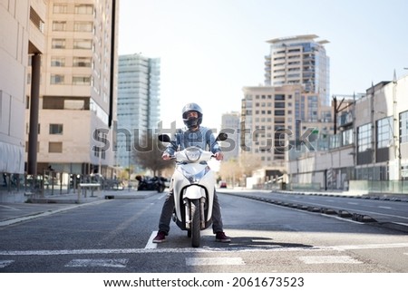 Wide shot of a young motorcyclist stopped at a traffic light in Barcelona. The man riding his scooter through the city on a large avenue lined with skyscrapers