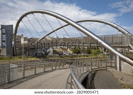A wide side view showcasing the dual sweeping steel arches and pedestrian ramps of the iconic Celtic Gateway bridge connecting Holyhead town centre to the port terminal in Anglesey, Wales. The modern architectural structure spans railway tracks and port infrastructure under a bright sky with light clouds.