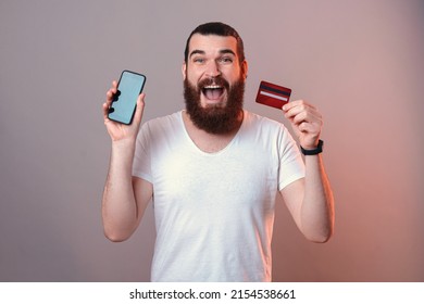 Wide Smiling Man Is Holding A Credit Card And Phone With Screen To The Camera. Studio Shot With Copy Space And Red Light Over Grey Background.