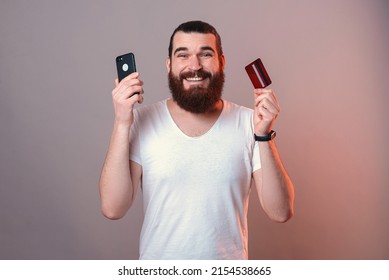 Wide Smiling Man Is Holding A Credit Card And Phone Near His Face. Studio Shot Over Grey Background With Red Light.