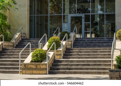 Wide Stairway With Metal Hand Railings And Small Shrubs Growing In The Flowerbed. It Goes Up To Entrance Of Multi Story Residential Building With Large Windows In Lobby.