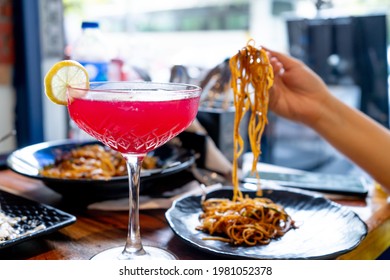 Wide Stemmed Wine Glass Filled With Red Pink Strawberry Cherry Bloody Mary Drink With Slice Of Lime With Out Of Focus Woman In Background Picking Up Noodles In A Fancy Restaurant Cafe In Delhi India