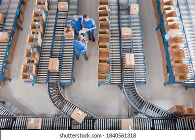 Wide Top View Of Workers With A Clipboard And Digital Tablet Having A Discussion Among Boxes Laid On Conveyor Belts At A Distribution Warehouse.