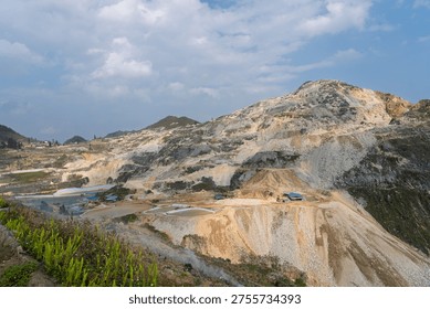 Wide view of an active tungsten mining site with terraced slopes, exposed earth, processing areas, and cloudy sky above a rugged mountainous environment.