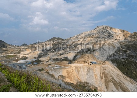 Wide view of an active tungsten mining site with terraced slopes, exposed earth, processing areas, and cloudy sky above a rugged mountainous environment.