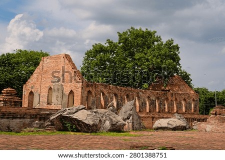 Wide view of ancient red brick ruins and large fallen stone debris at Wat Kudi Dao in Ayutthaya, Thailand. The historic temple hall features arched windows behind a paved courtyard and green trees