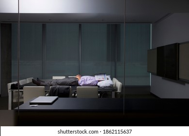 A wide view of a businessman in office clothes sleeping on a conference table with a pillow and a folder to cover his face