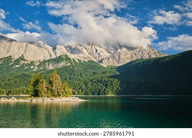 Wide view of the Eibsee lake surface with a solitary island and thick clouds covering the Zugspitze mountain peaks in Germany.