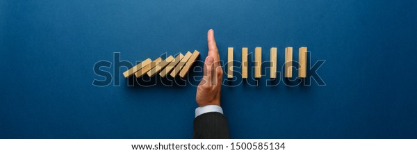Wide view image of businessman hand stopping\
collapsing dominos in a conceptual image. Top view over navy blue\
background.