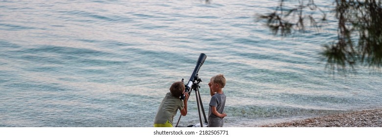 Wide View Image Of Two Boys, Brothers, Looking Through A Telescope Standing On Pebble Beach By The Sea.